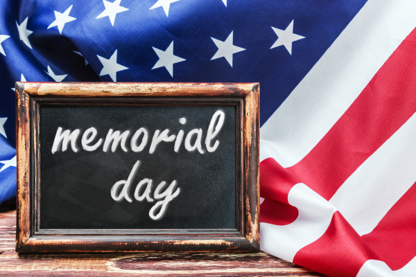 Chalkboard sign reading "memorial day" against an American flag, representing ADCO Hearing's Memorial Day sale on hearing accessibility devices